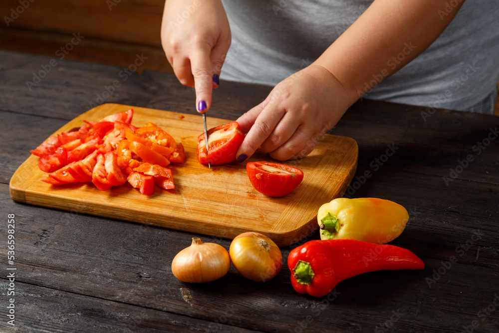 A woman in the kitchen with a knife cuts red tomatoes on a wooden board on the table next to onions and peppers. A woman in the kitchen with a knife cuts red tomatoes on a wooden board on the table next to onions and peppers.