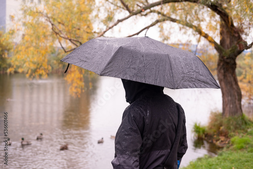 a man in a jacket with a hood, stands with his back with an umbrella in his hands, Against the background of the lake