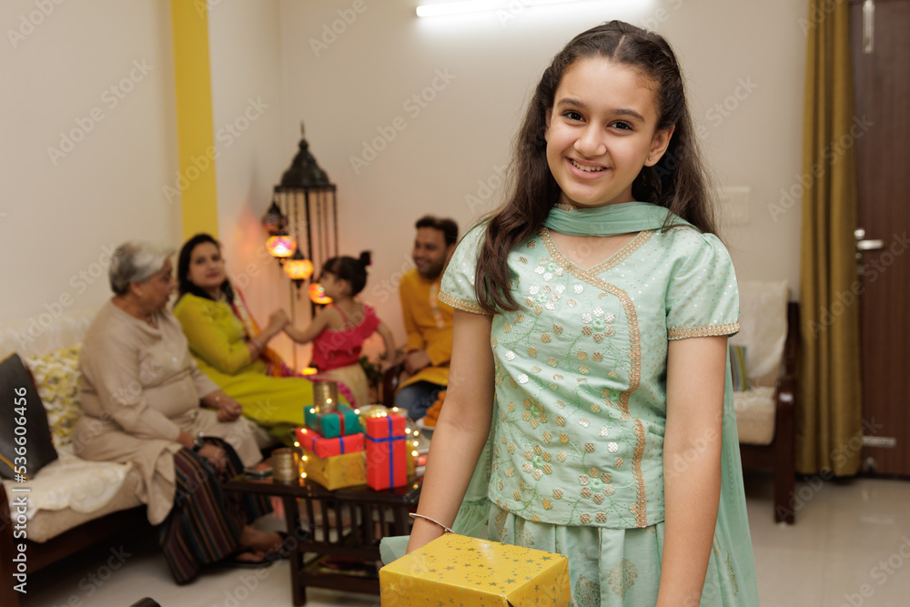 Fototapeta premium Young girl, kid, dressed up in ethnic wear holding gift boxes with smile expression pose for photo with family in background celebrating diwali Hindu festival Laxmi poojan