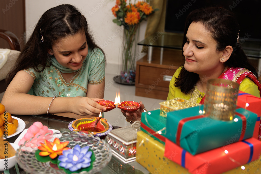 Fototapeta premium Mother and young daughter kid dressed up in ethnic wear, lighting up diya or lamp, celebrate diwali Hindu festival Laxmi poojan, kids expression