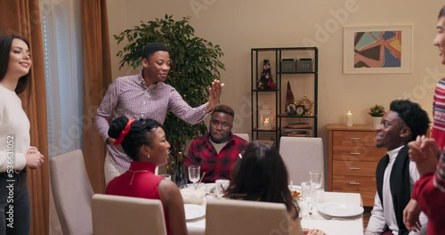 Man of African appearance invited friends to birthday party. Young people are sitting at the table, smiling and talking. His work colleagues entered the room and he introduced friends to each other.
