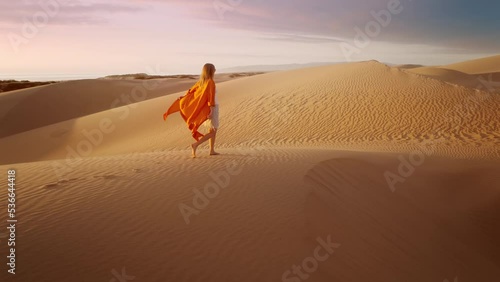 Barefoot female traveler in waving on wind orange dress walking by rippled sand desert surface with dark grey clouds at sunset. 4K Scenic nature aerial. Cinematic slow motion of woman hiking sand dune