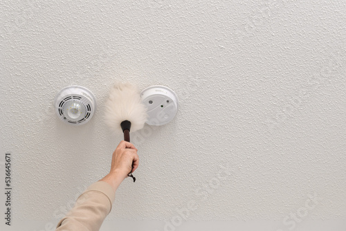 Canvas-taulu Woman reaching up to dust smoke alarm on ceiling at home