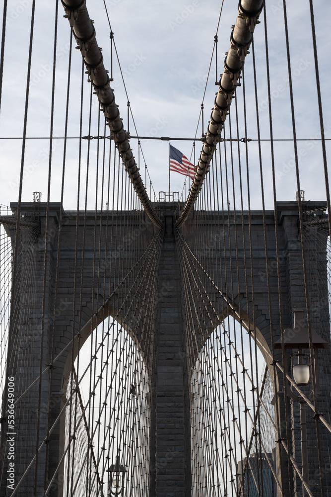 Fototapeta premium Cables leading up to a Brooklyn Bridge suspension tower, with an American flag on top