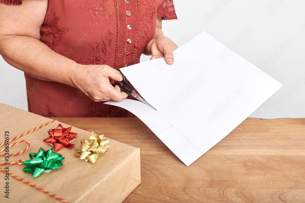 Unrecognizable mature woman cutting with scissors a piece of white cardboard, making handmade cards to put on her Christmas gifts. Simple design with copy space. Unrecognizable mature woman cutting with scissors a piece of white cardboard, making handmade cards to put on her Christmas gifts. Simple design with copy space.