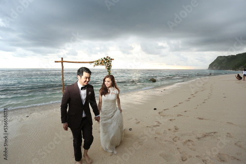 a man in suite and her woman in gown were holding hands  walking together along the beach during sunset