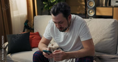 Happy guy sits on couch after waking up in pajamas. Man browses through phone, enters bank appication, high salary transferred to bank account, monthly bonus, joyful laughter. photo