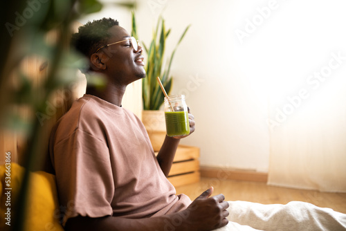 Relaxed black man drinking green smoothie at home. Side view of African American man enjoying healthy juice.