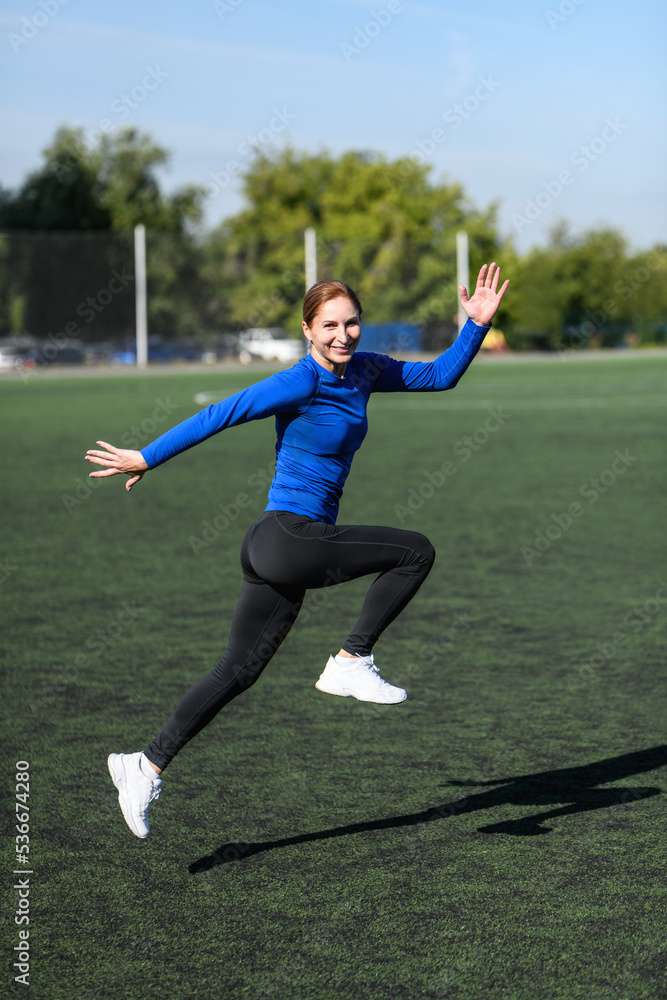Women and sport. Smiling Girl in sportswear - blue shirt and black leggings does exercises: bouncing and having fun on the grass at the stadium outdoor on a sunny day. Middle aged sportswoman Women and sport. Smiling Girl in sportswear - blue shirt and black leggings does exercises: bouncing and having fun on the grass at the stadium outdoor on a sunny day. Middle aged sportswoman