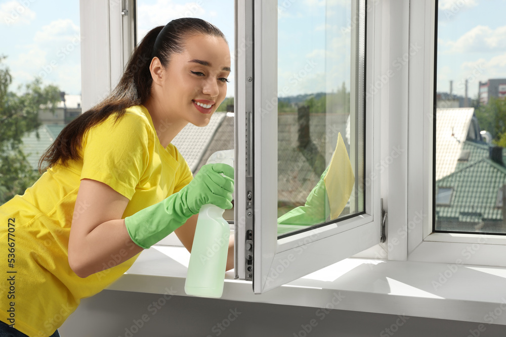 Happy young woman cleaning window glass with sponge cloth and spray indoors