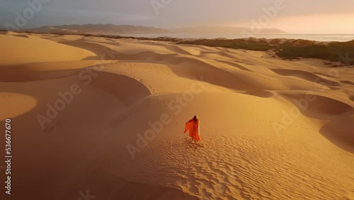 Woman in red orange dress standing on top of golden sand dune with raised hands while wind is blowing her dress and red hair. Freedom, nature concept 4k footage at cinematic golden sunset light on RED photo