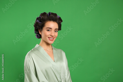 Happy young woman in silk bathrobe with hair curlers on green background, space for text © New Africa