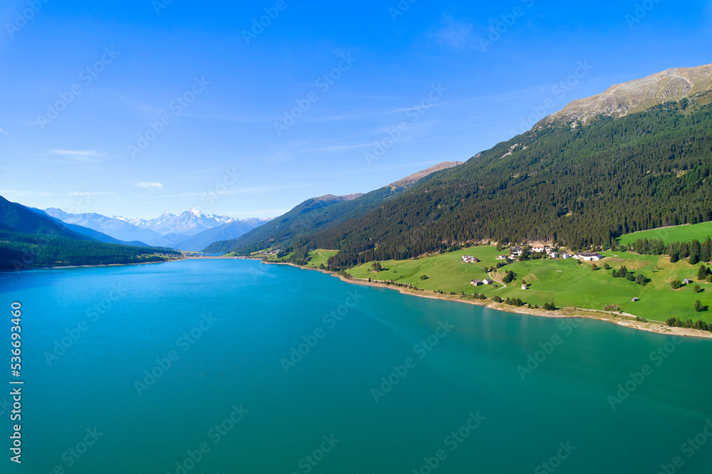 Aerial view of lake (Reschensee). Large reservoir surrounded by mountains at sunny noon. Recreation area for tourists and sportsmen. Organic farm on the shore. Italy, Vinschgau, Giern.