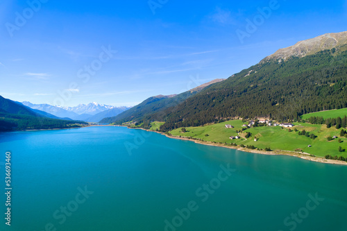 Aerial view of lake (Reschensee). Large reservoir surrounded by mountains at sunny noon. Recreation area for tourists and sportsmen. Organic farm on the shore. Italy, Vinschgau, Giern.