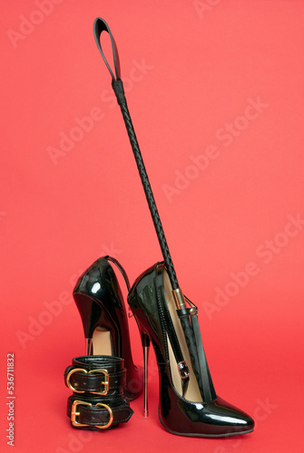 Adult sex games. Kinky lifestyle. Spank and a pair of black high-heeled shoes on the red background. Bdsm outfit