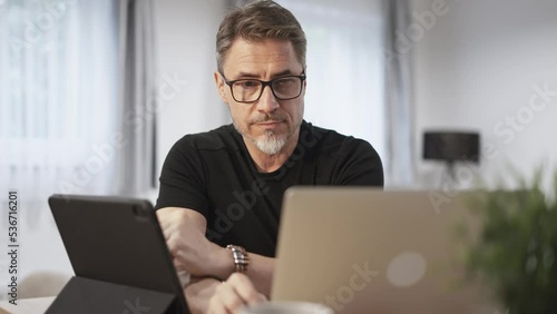 Older businessman working online with laptop and tablet computer at home sitting at desk in living room. Home office, browsing internet. Portrait of happy, mature age, middle age, mid adult man in 50s photo