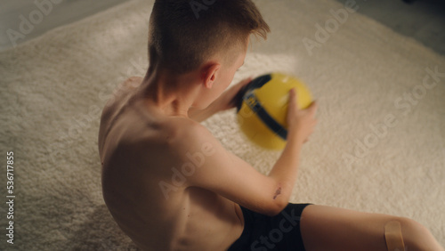 Young enthusiastic boy training abs holds a football while exercising alone indoors. Prepares before the soccer match. Sport and active lifestyle concept.