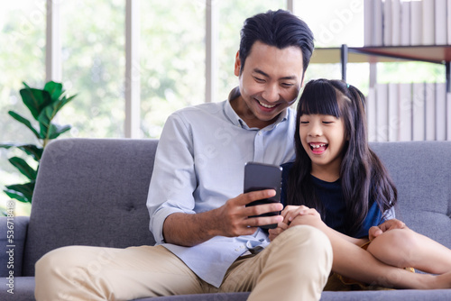 Young family smiling father and kid daughter sitting on couch watching playing smartphone together at home