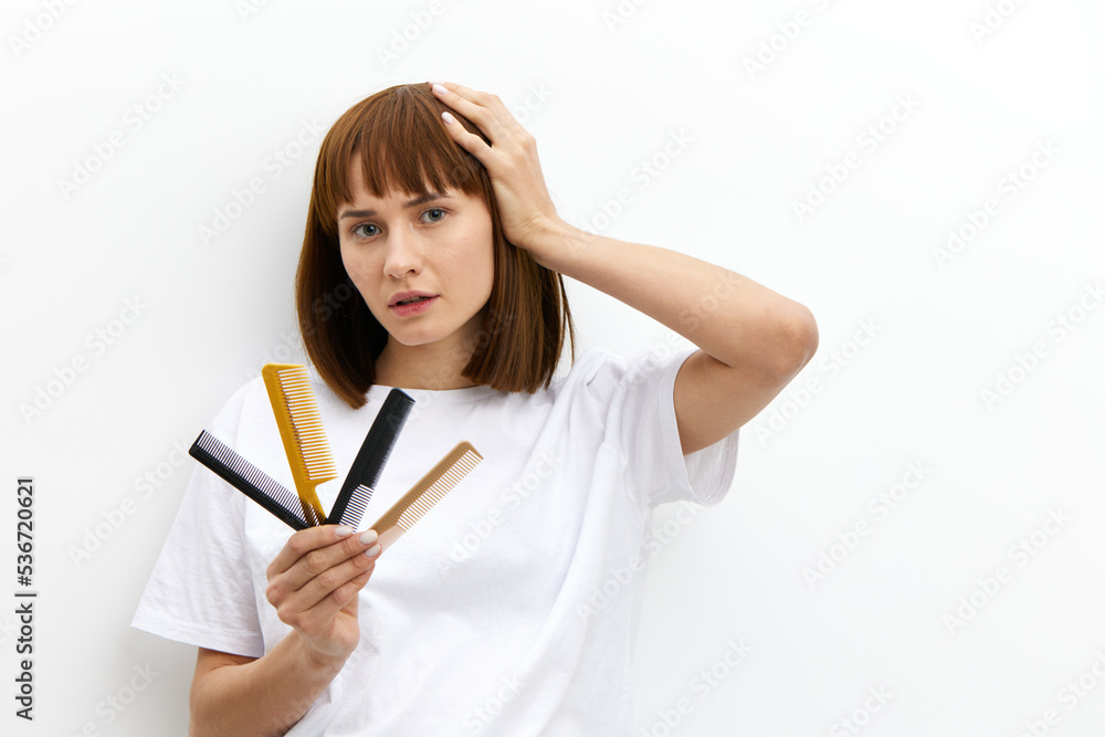 a sweet, pleasant woman with a beautiful hairstyle holds a lot of different combs in one hand, and touches her head with the other, standing in a white T-shirt on a white background a sweet, pleasant woman with a beautiful hairstyle holds a lot of different combs in one hand, and touches her head with the other, standing in a white T-shirt on a white background