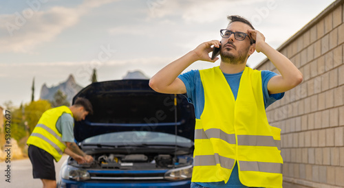 Fotografie Hombre con expresión de preocupación, llamando al servicio de asistencia por avería en el vehículo, con el técnico de fondo arreglando el coche