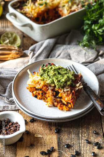 No meat enchiladas casserole dish with black beans, tomato, corn and sweet potatoes served on a plate on a wooden rustic table with guacamole dip on top