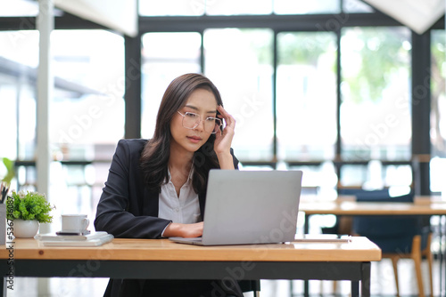 Exhausted businesswoman working at office desk with a laptop and touching her forehead, she is having an headache, stressful job concept