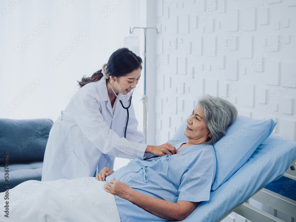 Fototapeta premium A beautiful young Asian woman doctor in white suit smiled while using stethoscope to examine, listen to heartbeat of elderly old female patient in blue dress who was lying on bed in hospital room.