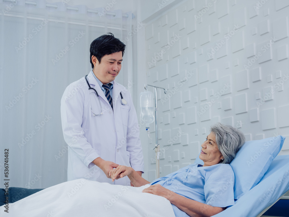 Obraz premium The kind Asian male doctor in white suit visits, talks and gives support, holding hand of happy elderly senior woman patient in light blue dress lying on bed in saline solution in hospital room.