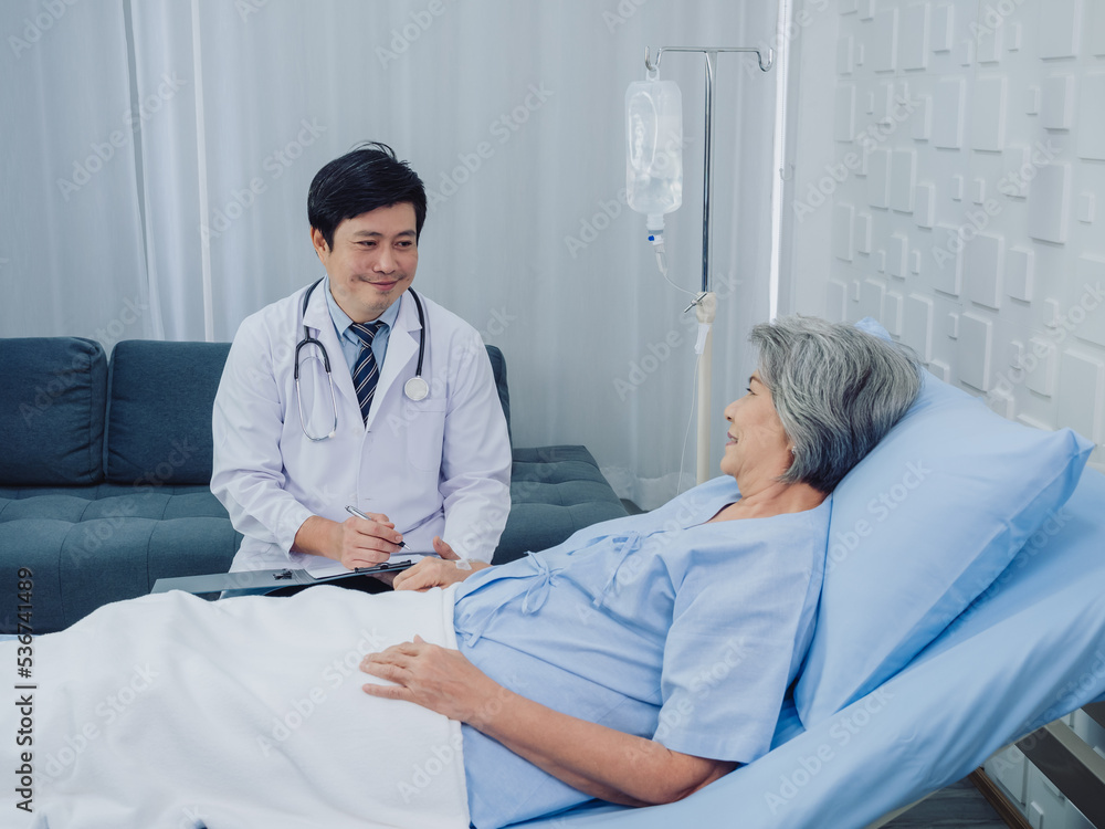 Obraz premium Smiling Asian doctor in white suit take notes while discussing happy Asian elderly, senior woman patient who lying on bed with receiving saline solution in hospital. Health care and medical concept.
