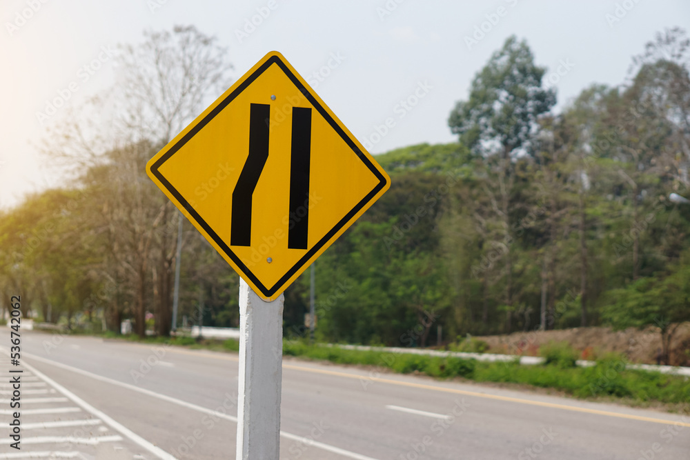 Yellow traffic square shaped left narrow lane sign at rural road Thailand to aware driver know Road narrows on left side ahead. Concept: Warning traffic sign for transportation. Yellow traffic square shaped left narrow lane sign at rural road Thailand to aware driver know Road narrows on left side ahead. Concept: Warning traffic sign for transportation.