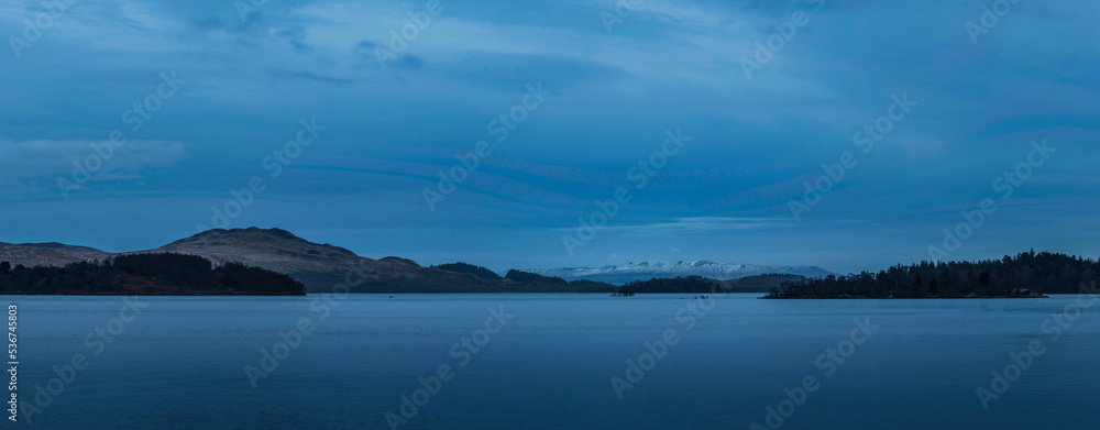 Beautiful landscape image of Loch Lomond and snowcapped mountain range in distance viewed from small village of Luss