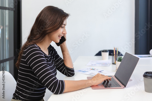 Smiling asian business woman talk on cell phone and using laptop. female freelance professional calling with customer work inquiries.