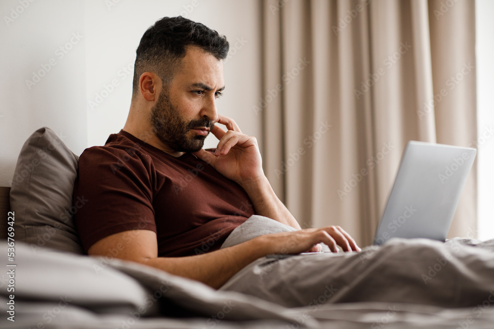 Smiling freelancer in home clothes working on modern laptop on comfortable bed at home, side view Smiling freelancer in home clothes working on modern laptop on comfortable bed at home, side view