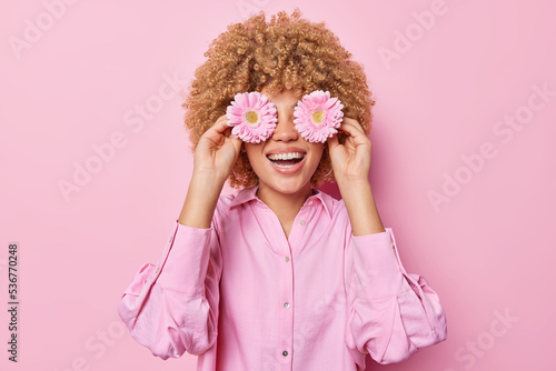 Happy curly haired young woman covers eyes with gerbera flowers smiles gladfully has fun dressed in fashionable blouse isolated over pink background. People florist and positive emotions concept