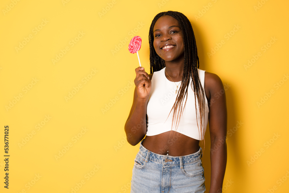 Smiling african girl holding candy lollipop. Young cheerful woman with lollipop isolated over yellow background, Smiling african girl holding candy lollipop. Young cheerful woman with lollipop isolated over yellow background,