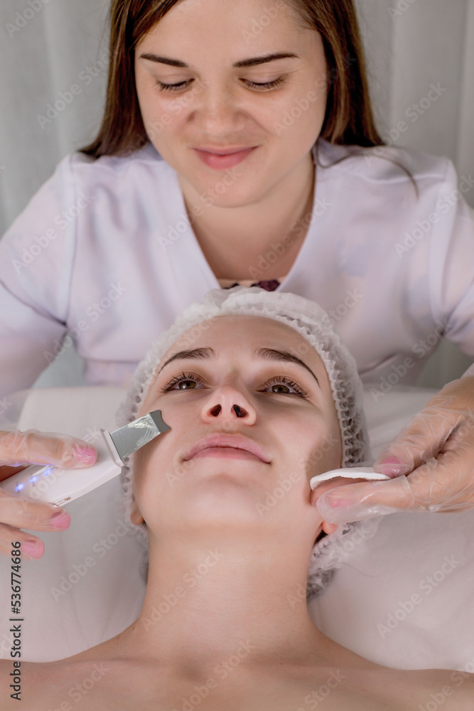 Naklejka premium Cosmetologist, beautician making facial treatment with ultrasonic spatula to young woman,face skin scrubber treatment with ultrasonic spatula,facial cleansing procedure in beauty salon