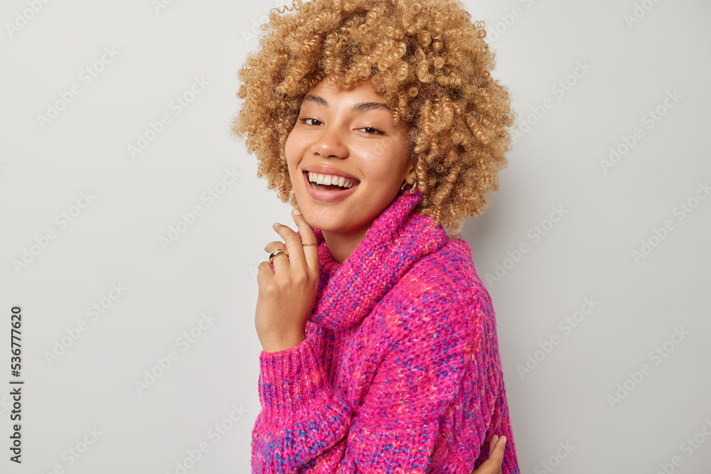 Beautiful young European woman with curly hair keeps hand under chin laughs positively dressed in pink knitted jumper smiles broadly isolated over grey background. Happy sincere emotions concept Beautiful young European woman with curly hair keeps hand under chin laughs positively dressed in pink knitted jumper smiles broadly isolated over grey background. Happy sincere emotions concept