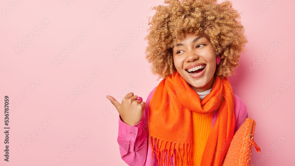 Positive carefree European woman with curly hair smiles broadly points thumb on left shows promo deal or advertisement wears orange scarf around neck carries bag isolated over pink background