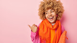 Positive carefree European woman with curly hair smiles broadly points thumb on left shows promo deal or advertisement wears orange scarf around neck carries bag isolated over pink background