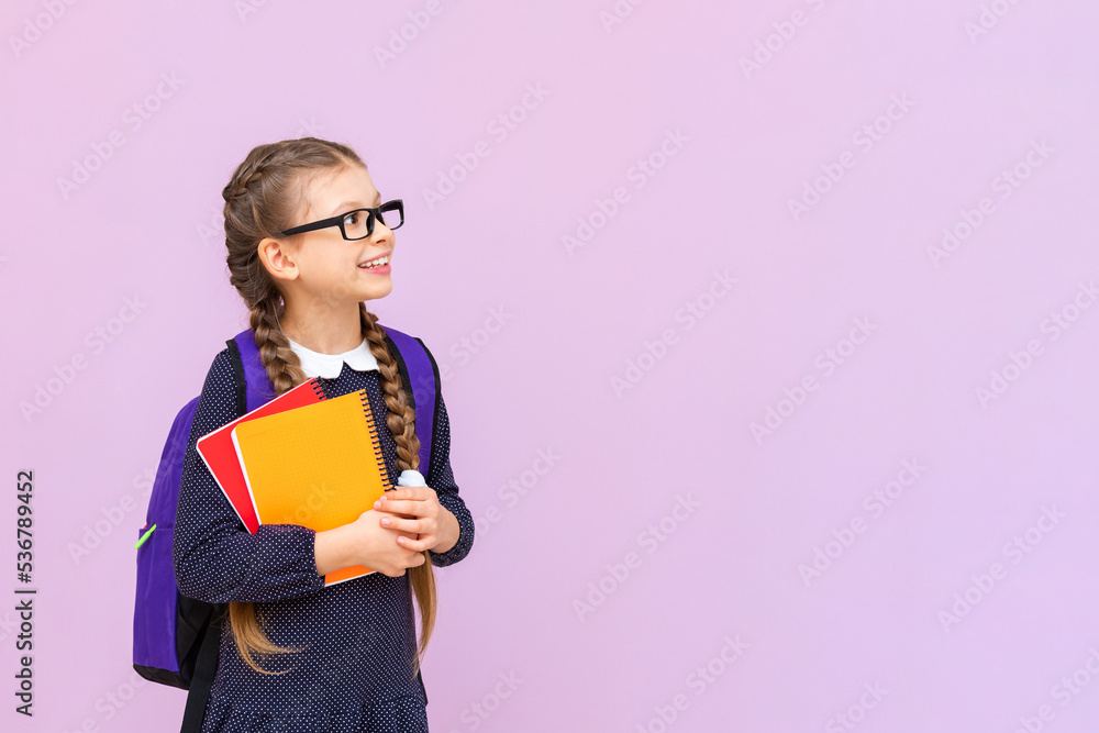 A little schoolgirl in glasses with a briefcase holds textbooks and looks at your advertisement on a pink isolated background. A little schoolgirl in glasses with a briefcase holds textbooks and looks at your advertisement on a pink isolated background.