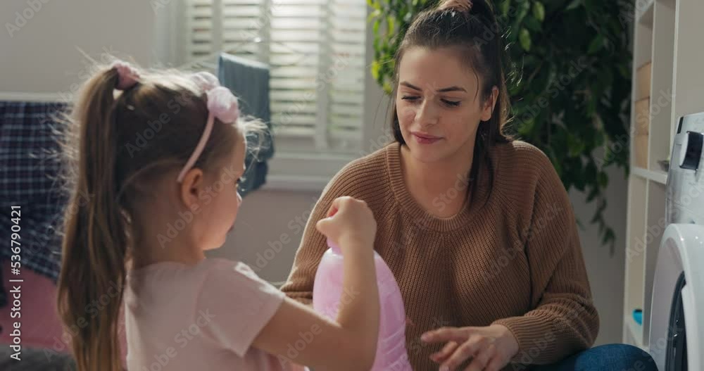 Young mother and daughter are doing laundry. Girl holds pink washing powder in hands and gives it to mother. Woman unscrews cork and smells aroma together with daughter. They are smiling and chatting. Young mother and daughter are doing laundry. Girl holds pink washing powder in hands and gives it to mother. Woman unscrews cork and smells aroma together with daughter. They are smiling and chatting.