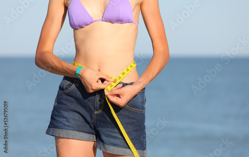 Canvas Print slender girl measures her waistline with yellow meter by the sea in summer weari