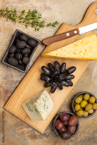 Two pieces of cheese and knife on the board. Green, red and black olives in bowls. Sprig of thyme and a bunch of black grapes