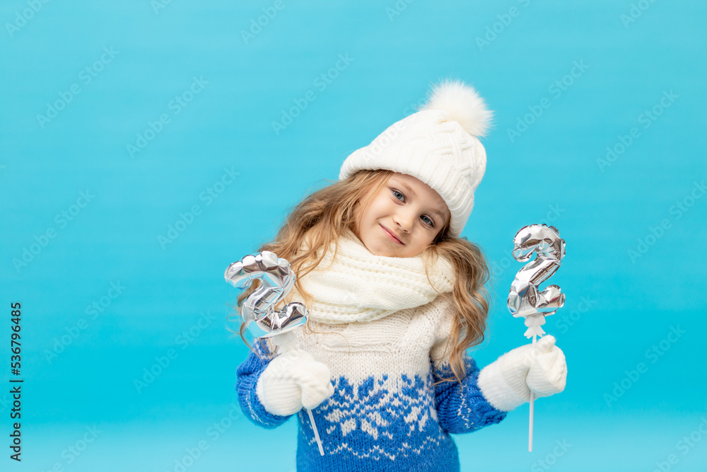 a little girl child on a blue background in the studio in a winter hat and sweater holds the numbers 2 and 2, the concept of new year and Christmas, space for text a little girl child on a blue background in the studio in a winter hat and sweater holds the numbers 2 and 2, the concept of new year and Christmas, space for text