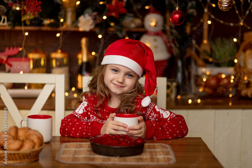 a cute little girl child in a red sweater and a Santa Claus hat is in the kitchen drinking tea with a pie and waiting for the new year or Christmas