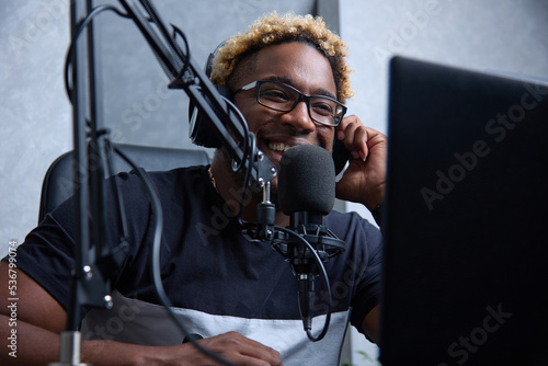 Fotografie A smiling cheerful black guy in headphones and with a professional microphone records an audio podcast for the radio