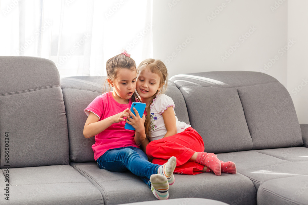 Stay Home Stay Safe. Two happy kids sitting on cozy chair relaxing playing in white living room indoors. Sisters having fun at home