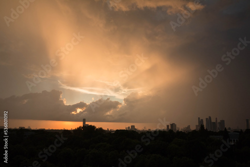 Panoramic view of the city at sunset. A skyscraper is visible in the distance. Beautiful sunset sky. Bright sun rays shine through gray clouds.