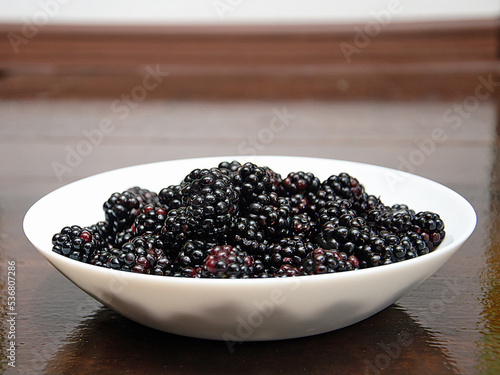 Black juicy blackberries on a white plate on a dark wooden background. blackberry berries