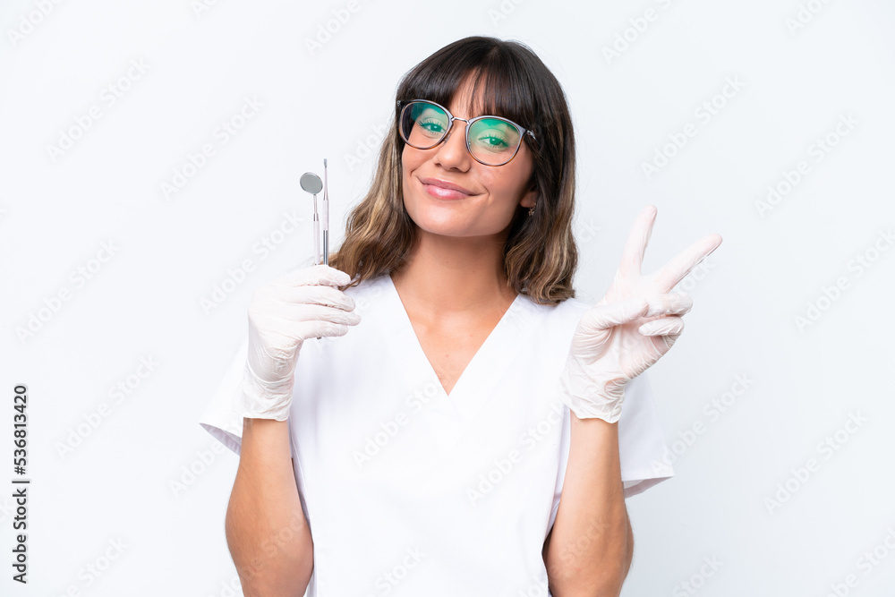 Dentist caucasian woman holding tools isolated on white background smiling and showing victory sign Dentist caucasian woman holding tools isolated on white background smiling and showing victory sign